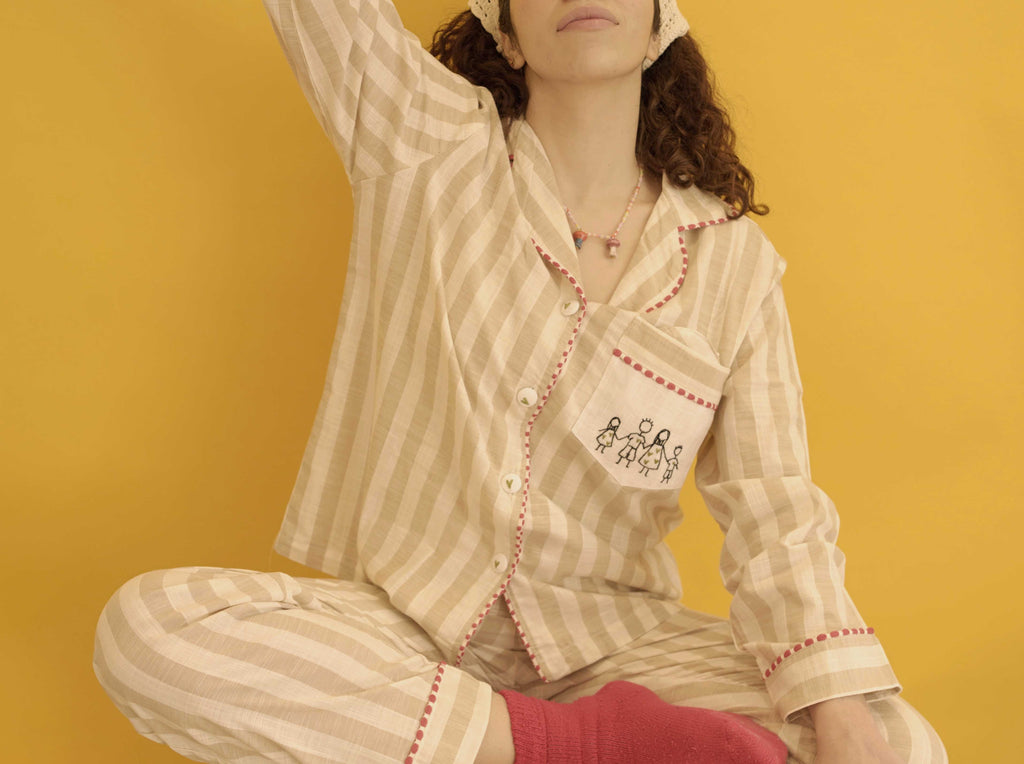 A model wearing a beige and white striped pajama set with red trim, sitting against a solid yellow background.