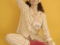 A model wearing a beige and white striped pajama set with red trim, sitting against a solid yellow background.