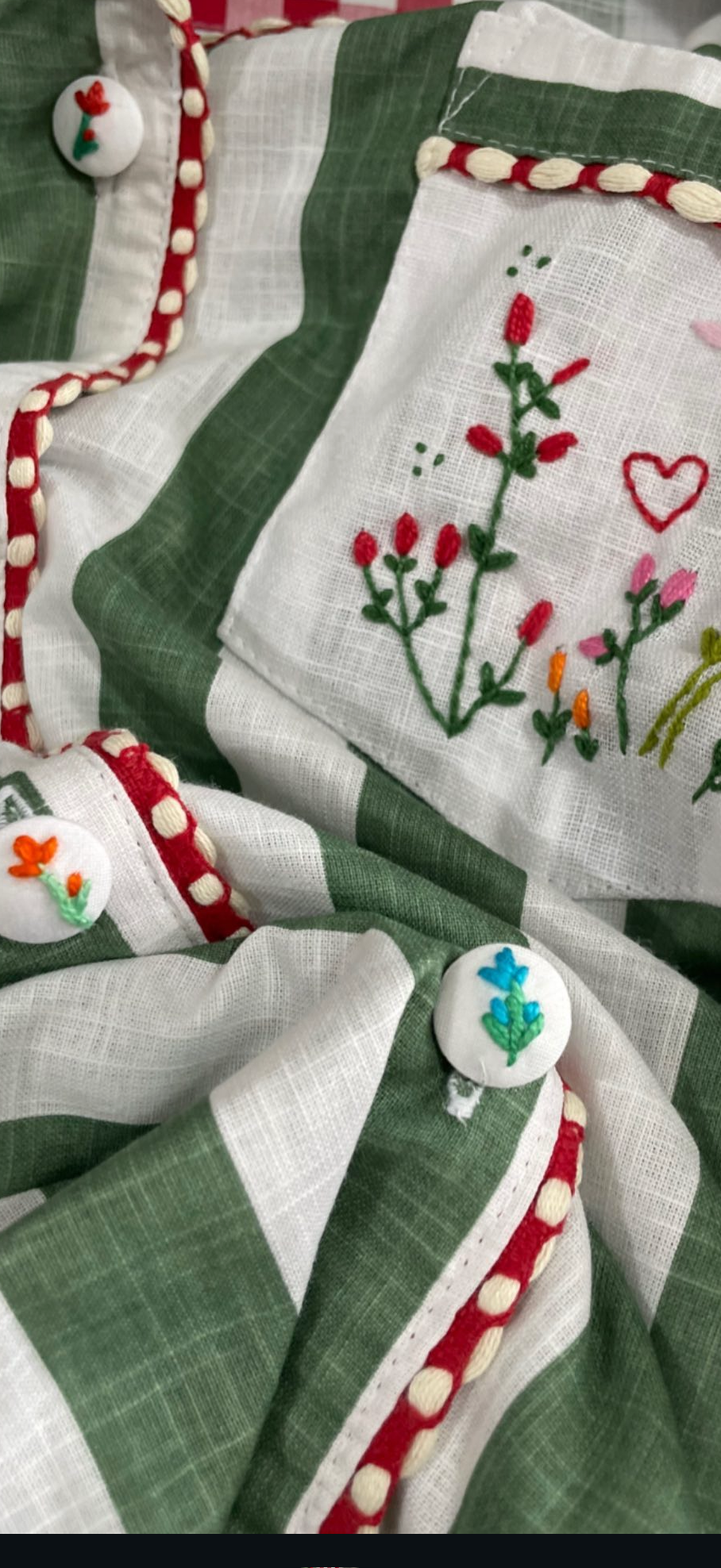 A close-up of a green and white striped fabric garment or textile with embroidered floral motifs, red trim, and decorative buttons, shown in a phone app screenshot with a navigation bar at the bottom.