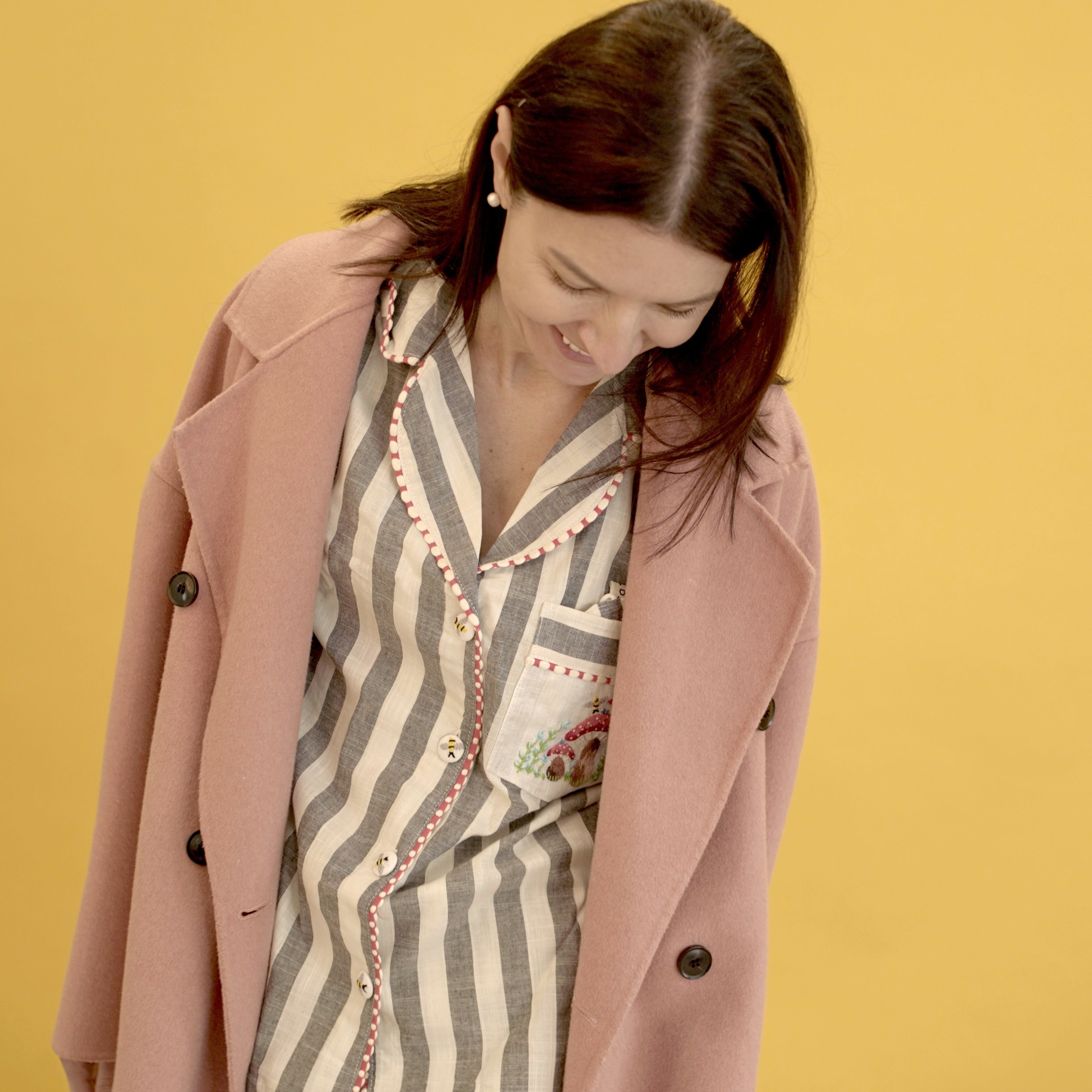 A woman wearing a light pink double-breasted coat over a beige and gray striped pajama top, standing against a solid yellow background.