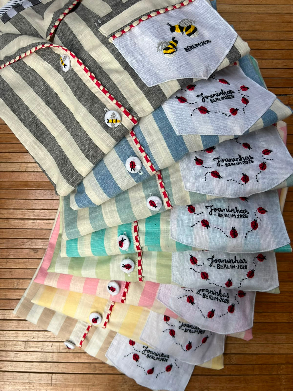 A stack of folded striped cotton PAJAMAS in various pastel and earthy tones, featuring embroidered details like bumblebees, ladybugs, and  random the text personalised names or images Johanna - Berlin 2025' on a wooden slatted surface. cotton poplin pyjamas. 