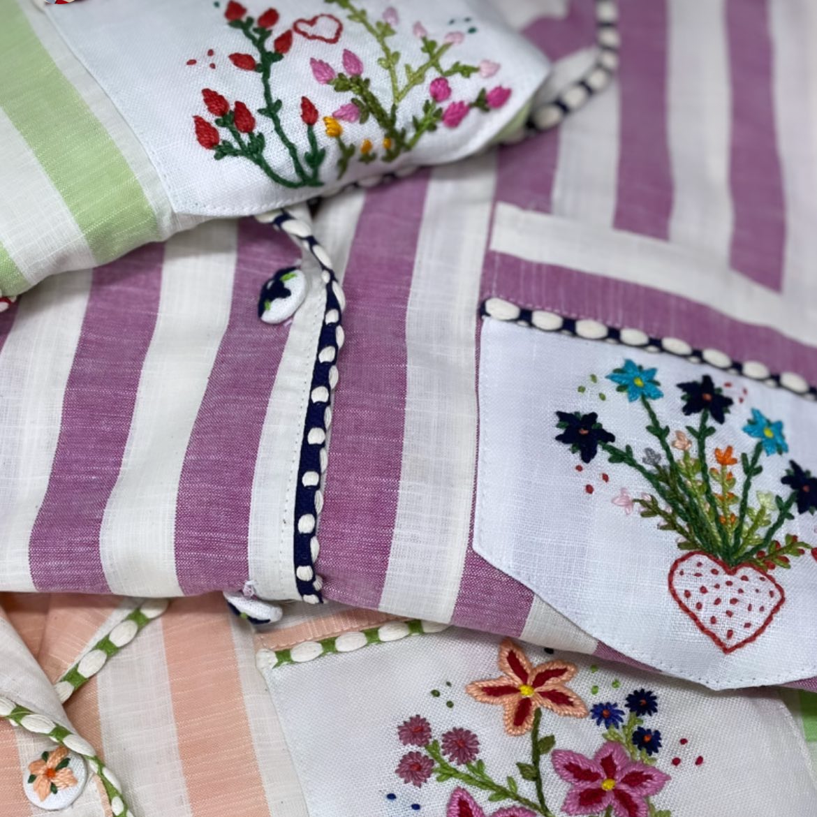 A close-up of folded striped fabric pieces with colorful floral embroidery, including purple, green, peach, and white stripes with embroidered flowers and leaves.